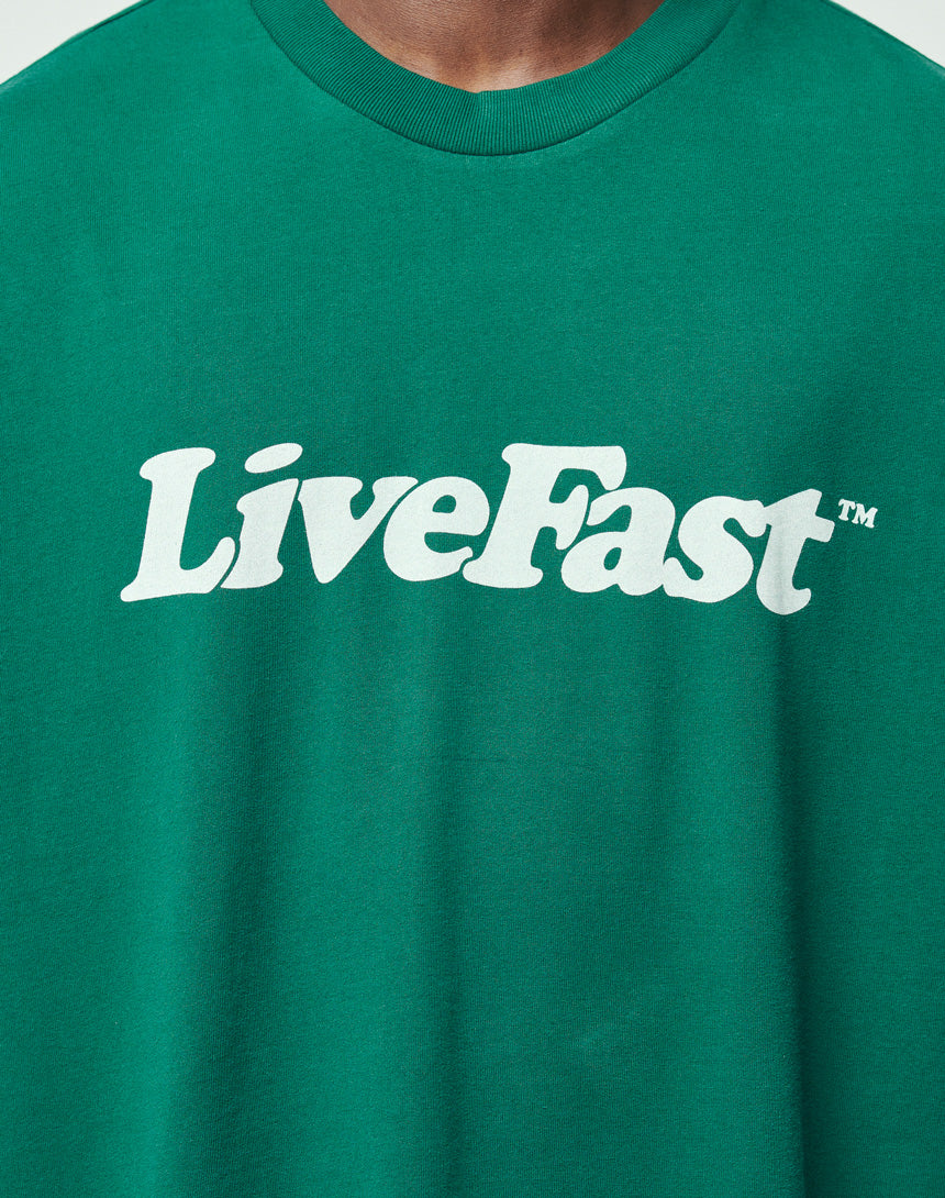 A person wears the LIVE FAST DIE YOUNG Basic Tee, a green regular fit shirt made from 100% Baumwolle, featuring a bold white LiveFast™ frontprint across the chest.