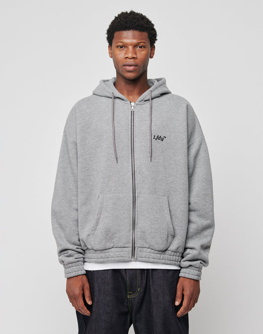 A young man faces the camera wearing a light gray LFDY Hooded Zip hoodie with doppellagige Kapuze and subtle logo-stick on the left chest, layered over a white shirt and dark jeans, against a plain light background.
