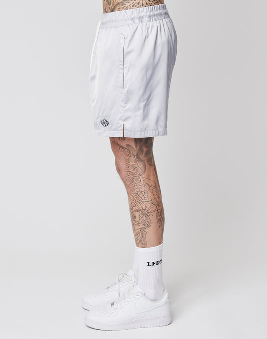 A person with leg tattoos is seen from the side wearing LIVE FAST DIE YOUNG Basic Satin Shorts, white socks with LEDY text, and white Nike sneakers, standing against a plain white background.