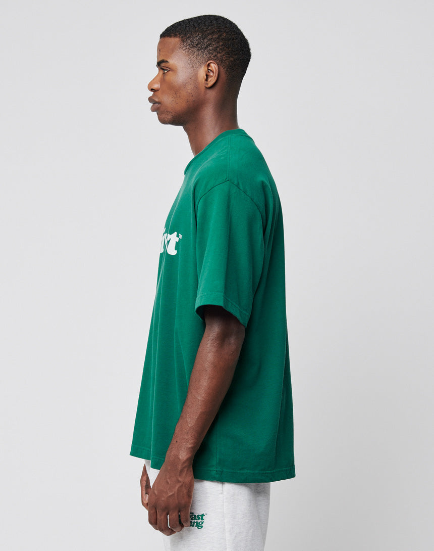 A man stands in profile facing left, wearing a loose-fitting green LIVE FAST DIE YOUNG Basic Tee with white frontprint text and light gray shorts. The background is plain and light-colored.