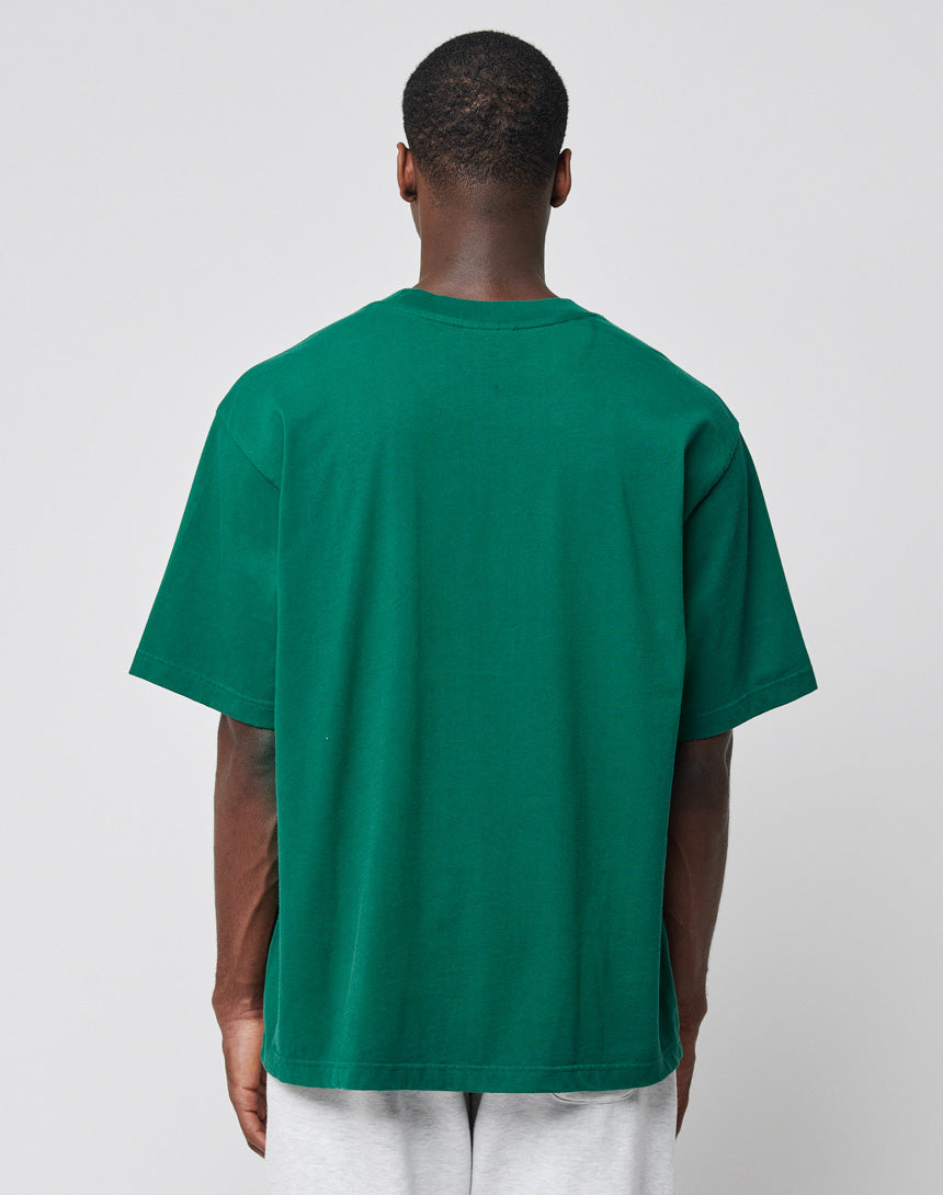 A person faces away from the camera, wearing the LIVE FAST DIE YOUNG Basic Tee—an oversized regular fit plain green T-shirt made from 100% Baumwolle—and light gray shorts, set against a plain light gray background.