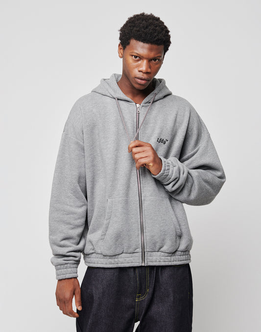 A young man with short curly hair wears the LFDY Hooded Zip hoodie in gray, featuring a doppellagige Kapuze and side pockets, paired with dark pants, against a plain light gray background. He holds the hoodie’s zipper with one hand.