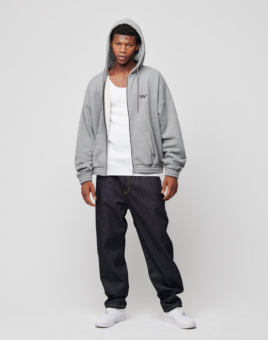 A young man stands against a plain light background, wearing an LFDY Hooded Zip gray hoodie with the hood up, a white tank top, dark baggy jeans, and white sneakers. His expression is neutral and his posture relaxed.