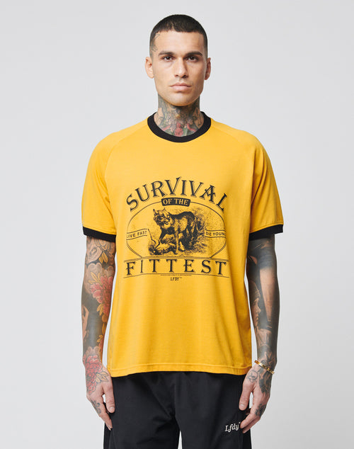 A tattooed person wears the LIVE FAST DIE YOUNG Survival Tee—a yellow relaxed fit shirt with black trim, raglan sleeves, Survival of the Fittest text, and a roaring tiger graphic—standing against a plain light background.