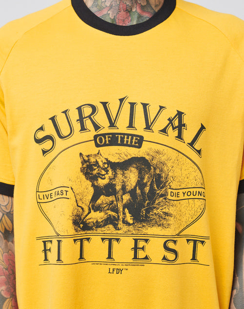 A person wears the LIVE FAST DIE YOUNG Survival Tee, a yellow cotton shirt with raglan sleeves, black trim, and wolf graphic. The text Survival of the Fittest; Live Fast, Die Young; LFDY appears as tattoos cover their neck and arms.