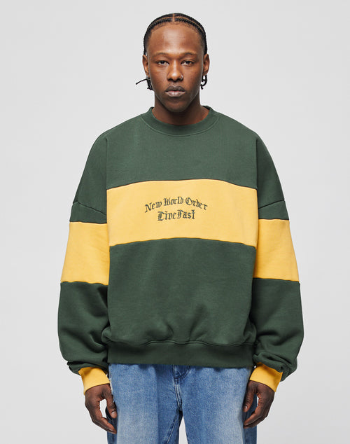 A man stands facing the camera in a green and yellow color-blocked New World Sweater by LIVE FAST DIE YOUNG, with New World Order Live Fast on the front. He has braided hair and wears blue contrast jeans against a plain light gray background.