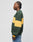 A man with braided hair stands in profile wearing an oversize fit green and yellow LIVE FAST DIE YOUNG New World Sweater made of cotton, paired with blue jeans against a plain light gray background.