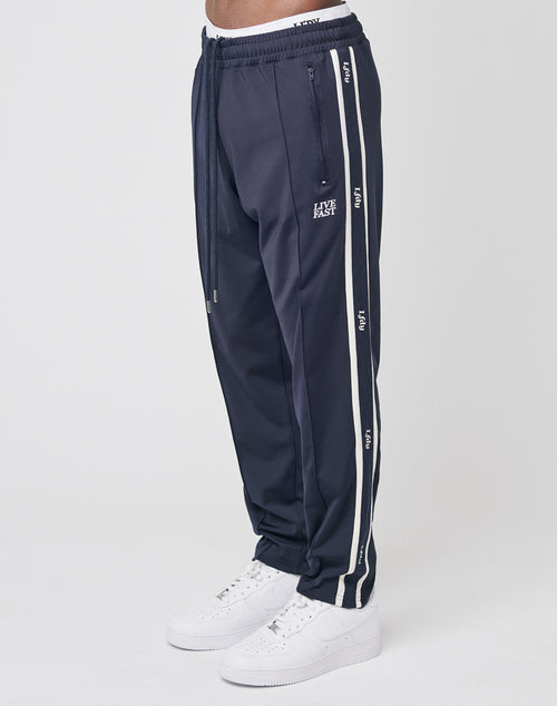 A person stands against a plain white background wearing navy blue LIVE FAST DIE YOUNG Trackjogger pants with white side stripes, Logostick detail, drawstring waist, and LAST LAST text near the pocket, paired with white sneakers.