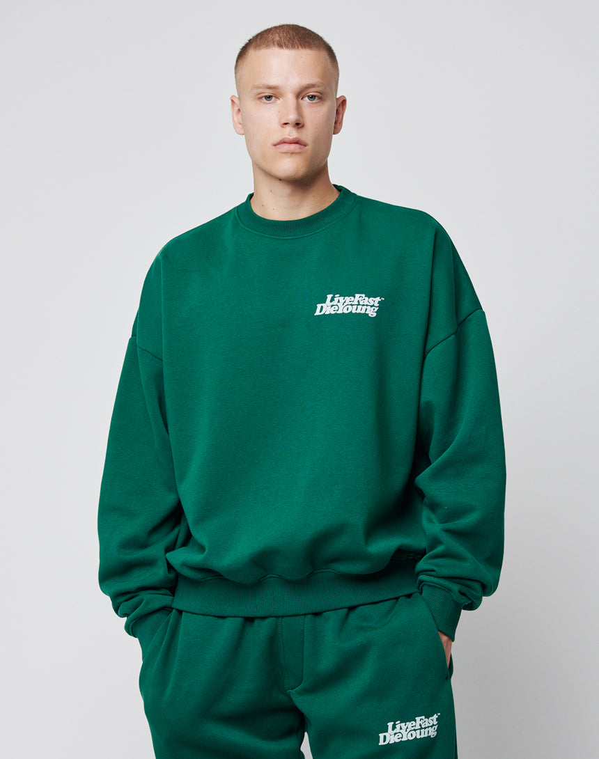 Wearing the LIVE FAST DIE YOUNG Basic Chest Sweater in dark green, paired with matching sweatpants featuring a bold chest print, a person stands against a plain light gray background.