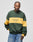 A person faces forward in a green and yellow New World Sweater by LIVE FAST DIE YOUNG, featuring Kontrastfarbe color-blocking and New World Order Live Fast text on the front, paired with blue jeans against a plain light background.