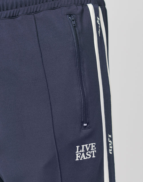 Close-up of the LIVE FAST DIE YOUNG Trackjogger in navy blue, featuring a white vertical stripe, Bügelfalte detail, zippered pocket, and LIVE FAST embroidery in white by the pocket.