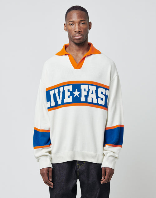 A man faces forward wearing the LFDY Knit Polo by LIVE FAST DIE YOUNG, an oversized white sweater with orange accents, blue stripes, and "LIVE FAST" boldly knit across the chest. The background is plain and light-colored.