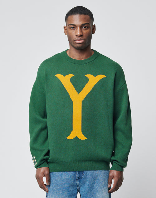 A man in a green LIVE FAST DIE YOUNG Y Knit Sweater featuring a bold yellow Intarsia-Logo letter Y on the front stands facing the camera with a neutral expression against a plain light gray background.