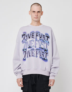 A person with a shaved head wears the LIVE FAST DIE YOUNG Triple Sweater—an oversized, light purple cotton sweatshirt with 7IVE FV5T printed three times in bold letters—paired with black pants against a plain white background.