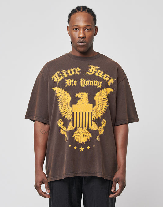 A man with braided hair and a neutral expression wears the LIVE FAST DIE YOUNG Eagle Tee, a loose brown regular-fit T-shirt with a large yellow eagle graphic and Live Fast Die Young text on 100% Baumwolle featuring a subtle vintage effect.