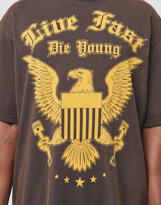 Eine Person trägt das Eagle Tee von LIVE FAST DIE YOUNG: ein Regular Fit T-Shirt aus 100% Baumwolle mit großem gelbem Adlerprint, Streifenschild, Live Fast Die Young-Schriftzug im Vintage-Look und drei Sternen unter dem Adler.