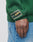 A person wearing blue jeans and a green LIVE FAST DIE YOUNG Y Knit Sweater in Oversize Fit, featuring an Intarsia-Logo and a 2023 Ten Years rectangular patch on the sleeve, stands with their hand relaxed at their side.