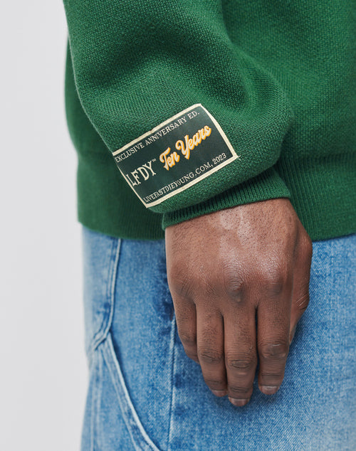 A person wearing blue jeans and a green LIVE FAST DIE YOUNG Y Knit Sweater in Oversize Fit, featuring an Intarsia-Logo and a 2023 Ten Years rectangular patch on the sleeve, stands with their hand relaxed at their side.