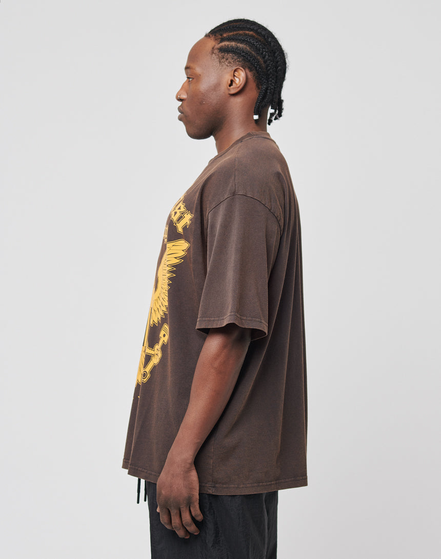 A man with braided hair stands in profile, wearing the LIVE FAST DIE YOUNG Eagle Tee— a loose brown regular-fit T-shirt with a yellow graphic— and black pants, set against a plain light gray background.