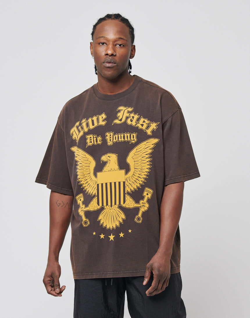 A man wearing the Eagle Tee by LIVE FAST DIE YOUNG, a regular fit vintage-style T-shirt with a large yellow eagle graphic and brand text, stands against a plain background. His braided hair and neutral expression highlight the 100% cotton look.