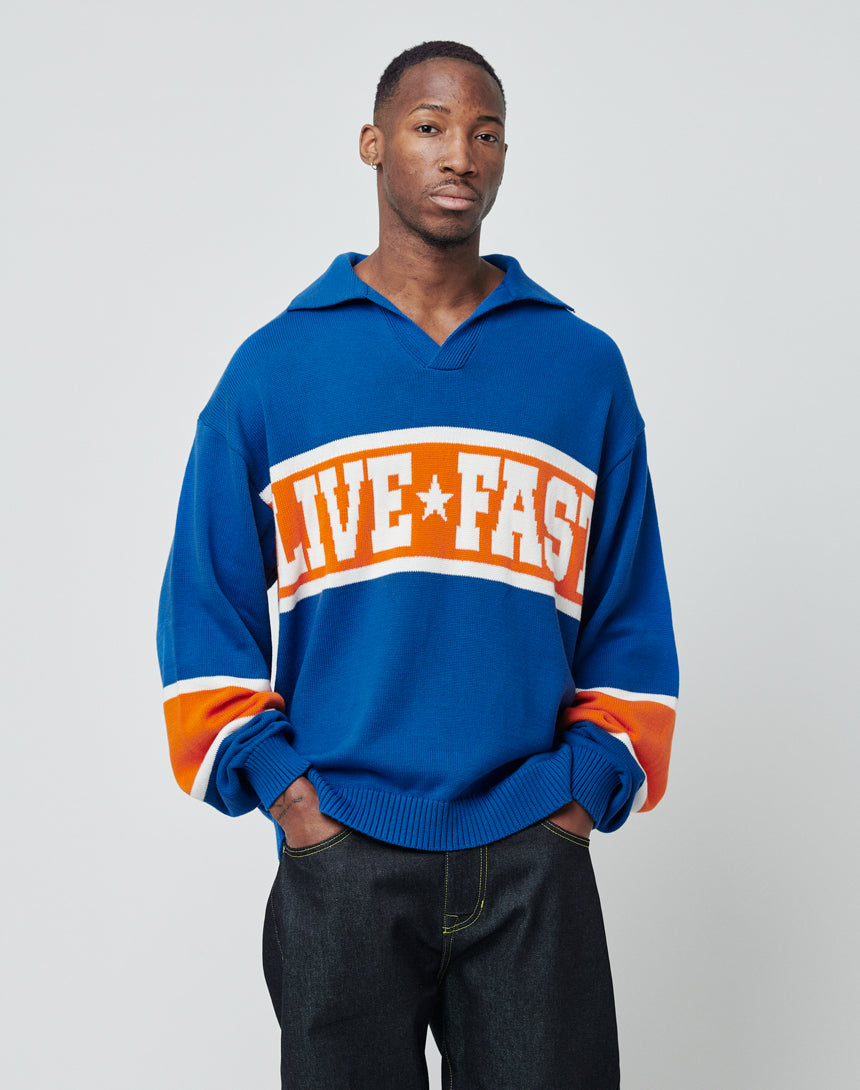 A man with a neutral expression stands against a plain light background, wearing the LIVE FAST DIE YOUNG LFDY Knit Polo. His hands are in his pockets as he models the knit polo.