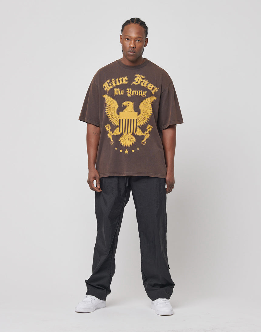 A man with braided hair stands against a plain background, wearing the Eagle Tee by LIVE FAST DIE YOUNG—a loose brown 100% cotton T-shirt featuring a gold eagle and vintage “Live Fast Die Young” text for a cool retro vibe.