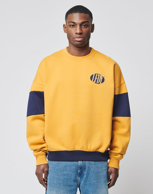 A man stands against a light gray background wearing the LFDY® Sweater by LIVE FAST DIE YOUNG—a yellow sweatshirt with navy blue sleeve stripes and a small circular logo on the left chest—paired with light blue jeans.