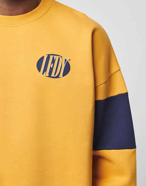 A person wearing the LFDY® Sweater by LIVE FAST DIE YOUNG: an oversize fit mustard yellow sweatshirt with a navy blue sleeve stripe, soft Baumwolle fabric, and an oval “LFDY” logo frontprint on the chest.