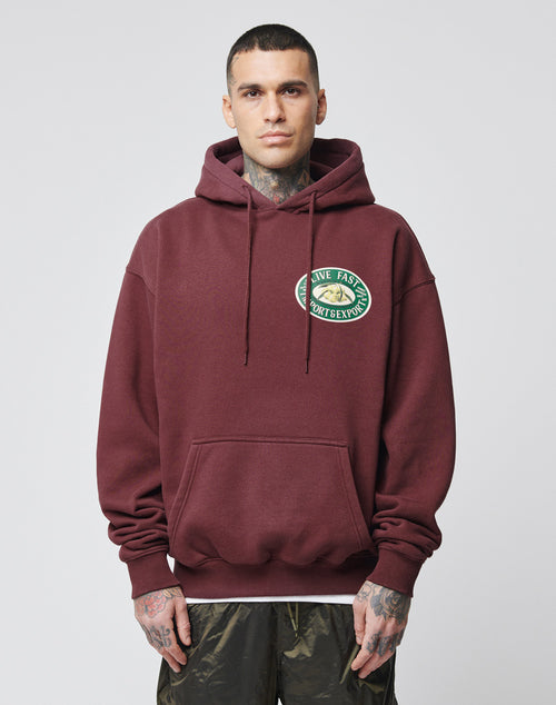 A person models the LIVE FAST DIE YOUNG Import & Export Hooded—a maroon oversized hoodie with a front pocket and a “LIVE FAST DONT EXPLOIT” patch—made from cotton-polyester, against a plain light gray background.