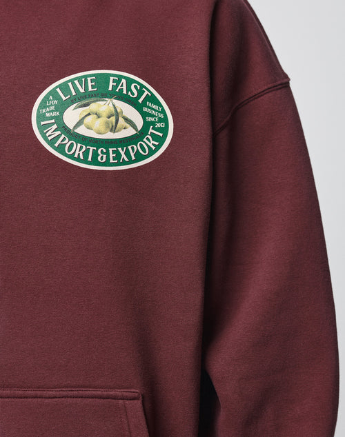 The Import & Export Hooded by LIVE FAST DIE YOUNG is a maroon oversized sweatshirt with a vintage effect, featuring a green and white chest patch reading “LIVE FAST IMPORT & EXPORT” with lemons and family business details since 2012.