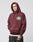 A man wears the LIVE FAST DIE YOUNG Import & Export Hooded sweatshirt, featuring a maroon color, green and white oval chest logo, and oversized fit, standing with one hand in his pocket against a plain light gray background.