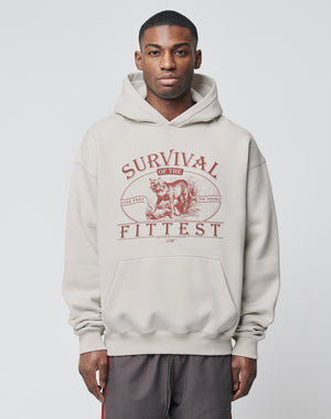 A young man stands facing forward in the LIVE FAST DIE YOUNG Survival Hooded, an oversized light gray cotton hoodie with a front pocket. A wolf graphic and Survival of the Fittest in red are on the chest. Easy care. Plain white background.