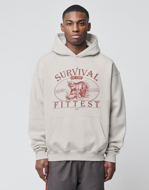 A young man stands facing forward in the LIVE FAST DIE YOUNG Survival Hooded, an oversized light gray cotton hoodie with a front pocket. A wolf graphic and Survival of the Fittest in red are on the chest. Easy care. Plain white background.
