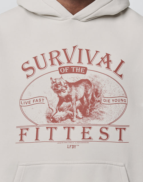 The cream-colored Survival Hooded by LIVE FAST DIE YOUNG features a red wolf graphic with SURVIVAL OF THE FITTEST and LIVE FAST DIE YOUNG text on the front; it’s oversized, made from cotton, and easy to care for.