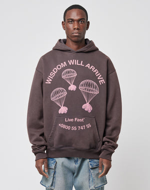 A man wears the LIVE FAST DIE YOUNG Wisdom Hooded—a dark, oversized cotton hoodie with pink parachute graphics and WISDOM WILL ARRIVE, Live Fast, and *0800 55 747 55 text, plus a vintage effect. He stands against a plain light background.