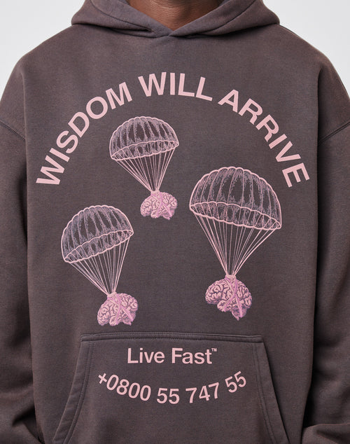 Wearing the LIVE FAST DIE YOUNG Wisdom Hooded, featuring pink brain-parachute graphics and “WISDOM WILL ARRIVE,” “Live Fast,” and +0800 55 747 55 text on the front. The oversize fit adds comfort and vintage style to the cotton fabric.
