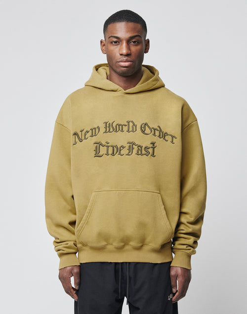 A man stands facing the camera in a light olive green New World Hooded by LIVE FAST DIE YOUNG, featuring black gothic New World Order Live Fast lettering. The Oversize Fit hoodie is crafted from premium Baumwolle and Polyester.