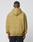 A person with short hair, seen from behind, wears the LIVE FAST DIE YOUNG New World Hooded oversize-fit tan hoodie (Baumwolle and polyester), hood down, paired with black pants against a plain light background.