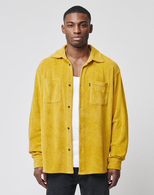 A man faces forward, wearing the LIVE FAST DIE YOUNG LFDY Frottee Shirt in yellow with an oversize fit and two-piece collar over a white shirt and black pants, set against a plain light gray background.