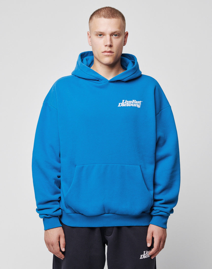 A person with short hair wears the bright blue LIVE FAST DIE YOUNG Basic Chest Hooded, featuring a front pocket and white chest print, while standing against a plain light background.