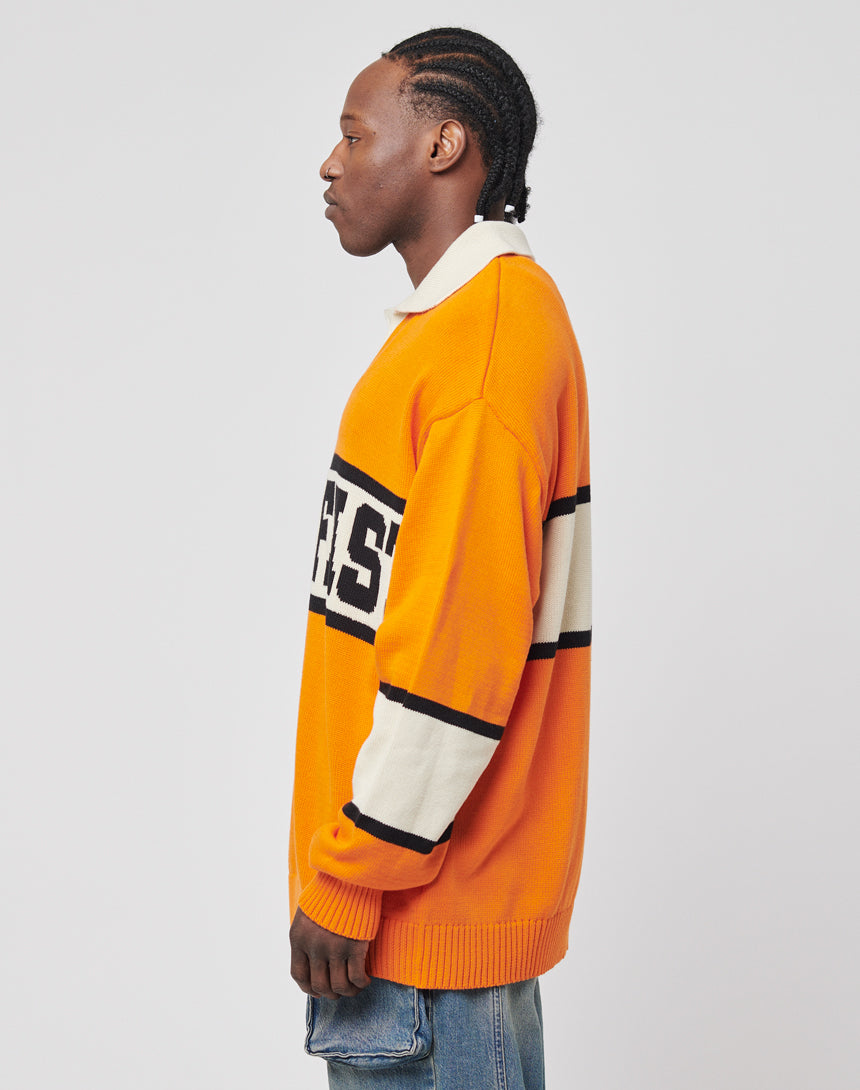 A man with braided hair stands in profile, wearing a LIVE FAST DIE YOUNG LFDY Knit Polo with black and white stripes and logo detail on the chest, paired with blue jeans, against a plain light background.