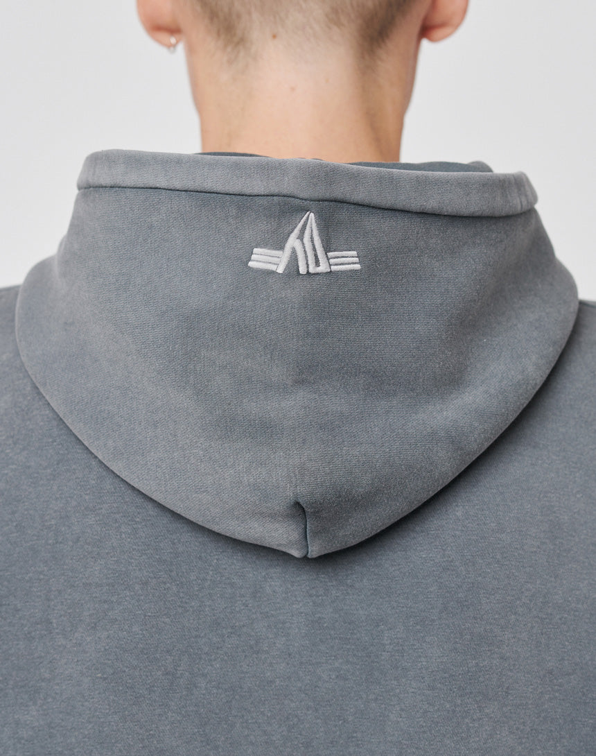 The image shows the back of a person wearing the LIVE FAST DIE YOUNG Triple LF Hooded sweatshirt in gray. It features a small white embroidered logo at the center of the hood and an oversized fit for a relaxed look. Short hair and ear are partly visible.
