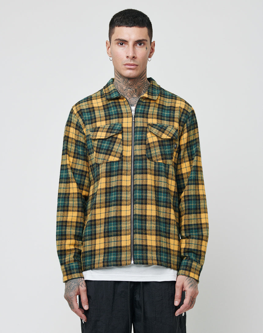 A person with short hair and neck and hand tattoos wears an LFDY Zip Flannel in yellow and green plaid over a white t-shirt, standing against a plain light background.