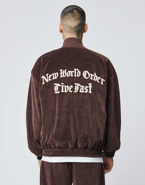 A person with short hair and neck tattoos, facing away from the camera, wears an oversize brown Velvet Tracksuit Jacket by LIVE FAST DIE YOUNG with New World Order Live Fast in large white gothic letters on the back.