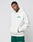 A man wears the LIVE FAST DIE YOUNG Basic Chest Hooded, an oversize white cotton hoodie with green text and a front pocket, paired with green pants. He stands with one hand in his pocket against a light grey background.