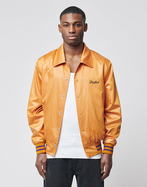 A man with short hair and a neutral expression models the LIVE FAST DIE YOUNG Satin College Jacket—an orange polyester button-up with striped cuffs, Loyalist embroidery, and a regular fit—against a plain light background.