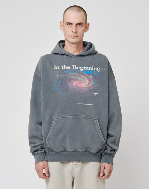 A person with a shaved head wears the LIVE FAST DIE YOUNG Beginning Hooded, a gray oversized hoodie featuring a bold galactic swirl frontprint with In the Beginning... text, standing against a plain white background.