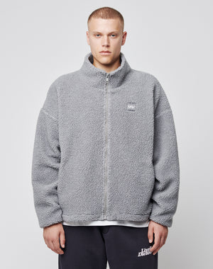 A person faces forward in a light grey Oversize Teddy Jacket by LIVE FAST DIE YOUNG, featuring an oversize fit and small chest logo-patch, worn with dark pants against a plain white background.