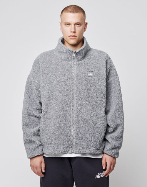 A person faces forward in a light grey Oversize Teddy Jacket by LIVE FAST DIE YOUNG, featuring an oversize fit and small chest logo-patch, worn with dark pants against a plain white background.