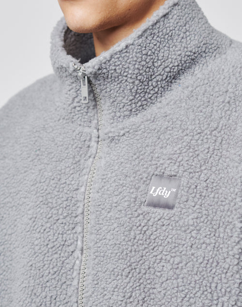 Close-up of a person wearing the Oversize Teddy Jacket by LIVE FAST DIE YOUNG, in light gray. The textured jacket has a high collar, visible zipper, oversize fit, and a small rectangular Lftdy™ logo patch on the chest.